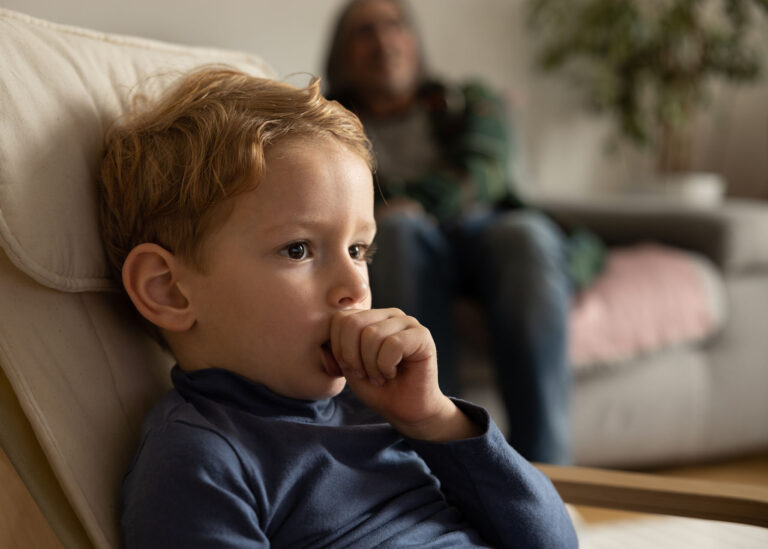 A little boy sits at home in a chair and sucks his finger, in the background a man watches the boy